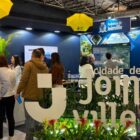 Homens e mulheres visitam um estande em uma feira. No local, há paineis azuis, duas televisões, alguns guarda-chuvas amarelos e um painel decorativo escrito joinville com flores