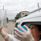 Mulher vestida com equipamento de proteção está no banco traseiro de um carro. A imagem externa ao veículo mostra a mulher com os braços para fora, com uma das mãos segurando um frasco de plástico de onde saem mosquitos