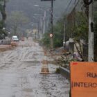 Imagem mostra rua de chão em dia de chuva. Há uma placa laranja informando que a rua está interditada