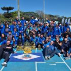 Dezenas de pessoas com uniformes azuis posam para foto em uma quadra de piso azul. Há uma bandeira de Joinville e vários troféus no chão
