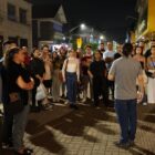 Imagem mostra uma rua de Joinville à noite. Um homem de camisa cinza está de costas para a câmera, falando com vários homens e mulheres que estão em pé, escutando o que ele diz