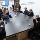 Imagem mostra uma sala com grande mesa em que há vários homens e mulheres sentados em ambos os lados e uma mulher em uma das pontas. Na parede oposta, há uma televisão que exibe uma tela escrito estradas rurais