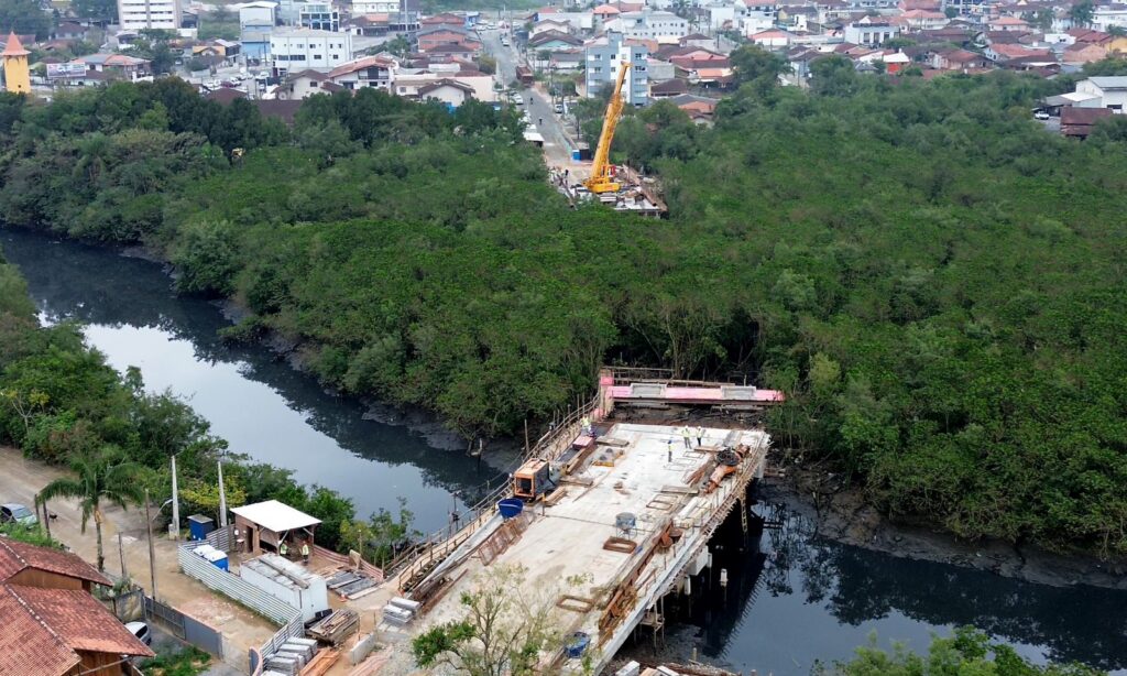 Imagem aérea mostra obras da Ponte Anêmonas. Há estruturas construídas em ambos os lados de uma área de vegetação por onde passa um rio. No entorno desta área, há ruas e residências.