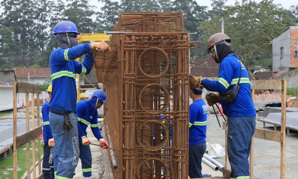 Quatro trabalhadores com uniformes azuis e capacetes manuseiam uma grande grade feita de ferro, usada para concretagem
