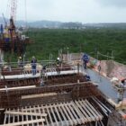 Imagem aérea mostra construção da Ponte Joinville, com trabalhadores em área de ferro e concreto