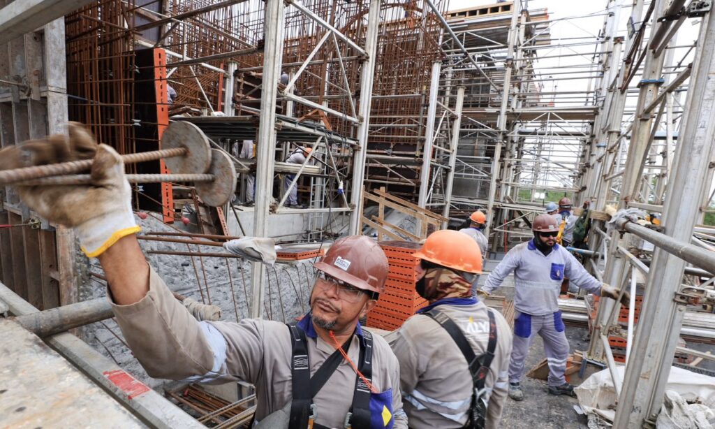 Nas obras de construção da Ponte Joinville, há no primeiro plano um trabalhador de capacete manuseando barras de ferro. No fundo, há outros homens uniformizados trabalhando