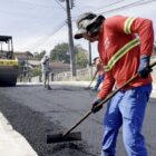 Homem com uniforme está segurando uma espécie de pá com a qual nivela o asfalto na rua. Ao fundo, há um rolo compressor também fazendo a pavimentação