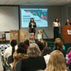 Três mulheres estão à frente de uma sala ampla, em um palco. Uma delas fala ao microfone para uma plateia onde há pessoas sentadas. Outra mulher está se comunicando em libras e a terceira está parada atrás de um púlpito