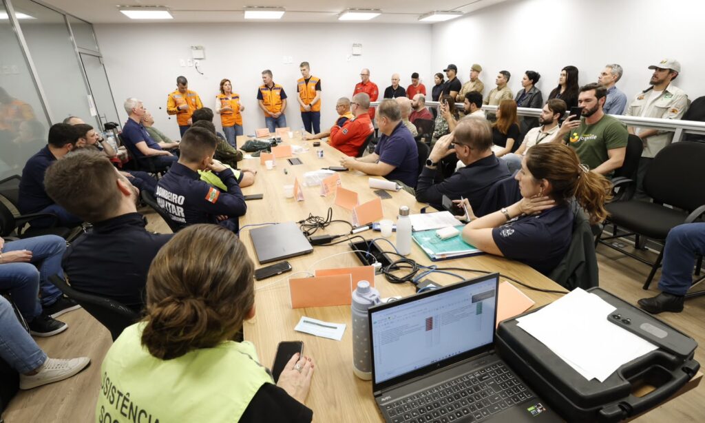 Imagem mostra uma sala ampla com uma grande mesa no centro e dezenas de homens e mulheres sentados e em pé. Na cabeceira da mesa, há três homens e uma mulher em pé, vestidos com colete laranja