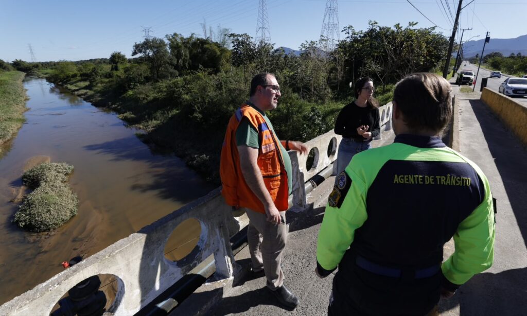 Homem com colete laranja e duas mulheres estão em uma ponte com um rio com baixo volume de água ao fundo