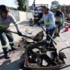 Três trabalhadores uniformizados estão fazendo a manutenção no asfalto de uma rua. Em primeiro plano, dois deles estão com pás, próximos à área de reparo. Ao fundo, o outro homem manuseia um equipamento perfurante