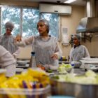 Adolescentes estão vestidos com aventais e tocas em uma cozinha. No centro da imagem, uma garota segura um pote. Em primeiro plano, há potes com alface e outros alimentos