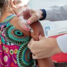 Imagem mostra os braços de uma pessoa usando jaleco segurando o braço de uma menina criança. Uma das mãos segura o braço enquanto a outra aplica uma vacina