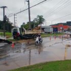 Imagem mostra rua pavimentada com água sobre a pista no momento em que passa uma moto pelo local. No trecho, há dois caminhões e cones sinalizando a pista