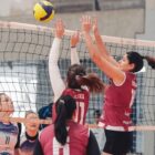 Em uma quadra de vôlei, três jogadoras aparecem de um lado da rede e duas estão de outro. Elas se movimentam para atingir a bola, que está acima da rede.