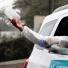 Homem vestido com equipamento de proteção está no banco traseiro de um carro. A imagem externa ao veículo mostra o homem com os braços para fora, com uma das mãos segurando um frasco de plástico