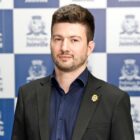 Homem branco de cabelo castanho usa camisa social e blazer e posa para a câmera em frente a um painel da prefeitura de joinville