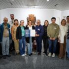 Dez pessoas, entre homens e mulheres, estão em pé, posando para a câmera. No centro um homem e uma mulher exibem um documento. Todos sorriem