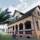Imagem mostra a Casa Krüger, uma casa histórica amarela com detalhes em vermelho, floreiras na varanda e jardim na frente
