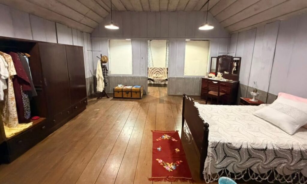 Em um cômodo com forro, paredes e piso de madeira, há uma cama, um guarda-roupas, cômoda e outros móveis antigos