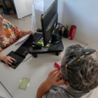 A foto tirada de cima mostra uma mulher atendendo um homem em um balcão com computador e outros objetos de informática. A mulher sorri
