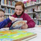 Mulher ruiva e menino vestido com fantasia de herói sorriem enquanto folheiam um livro infantil em cima de uma mesa