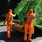 Dois homens uniformizados de laranja estão próximos a um caminhão da coleta de lixo