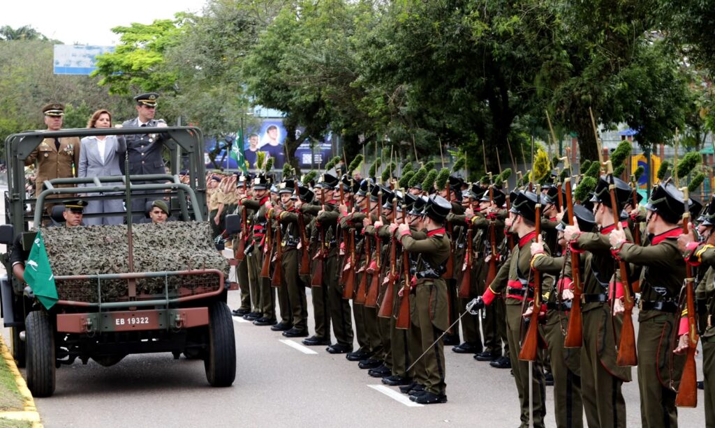 Jipe com dois comandantes e a prefeita passa ao lado de um pelotão do exército, com integrantes que seguram armas