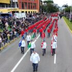 Imagem aérea mostra integrantes de um pelotão vestidos de vermelho e cinza desfilando na avenida Beira Rio. Ao lado da rua, o público acompanha o desfile