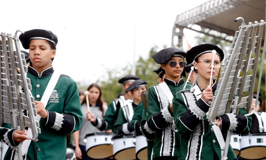 Adolescentes uniformizados com roupas verdes tocam instrumentos na fanfarra enquanto desfilam