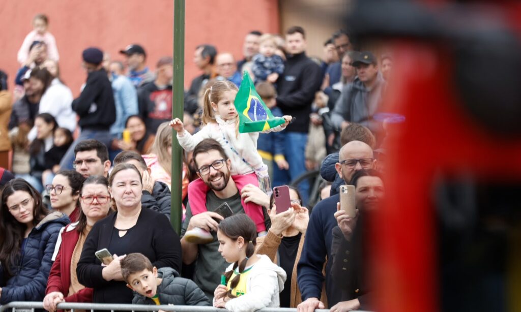 Público acompanha desfile de 7 de setembro em Joinville. Uma menina sentada no pescoço do pai segura uma bandeira do Brasil