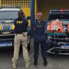 Duas mulheres, uma com uniforme da PRF e outra da Guarda Municipal, estão de costas para a câmera. Ao lado delas, há duas viaturas, uma com vários tabletes de carro no capô