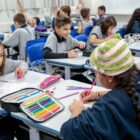 Em uma sala de aula, alunos e alunas adolescentes estão uniformizados, sentados em cadeiras, com o material escolar sobre as mesas. Eles escrevem, desenham e alguns sorriem