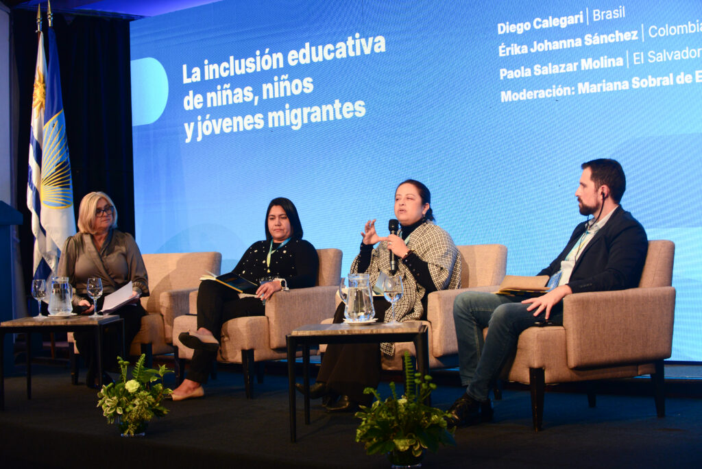 Três mulheres e um homem estão sentados em poltronas em um palco. Atrás deles há um grande painel de LED azul com escrita em espanhol