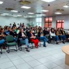 Imagem mostra uma ampla sala com várias pessoas, entre homens e mulheres, sentadas em cadeiras