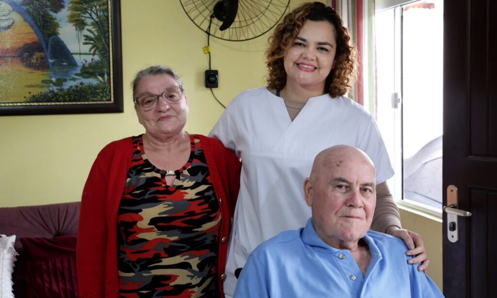 Mulher idosa de casaco vermelho e mulher jovem de cabelos encaracolados e jaleco branco estão em pé, ao lado de um homem idoso careca, que está sentado. Todos sorriem para a foto.