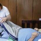 Mulher com jaleco branco e cabelo encaracolado está tocando as pernas um homem idoso que está deitado sobre uma cama. Ela está pressionando um dos pés dele, enquanto puxa uma de suas pernas.