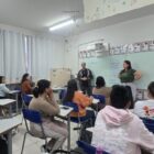 Imagem mostra uma sala de aula com vários adultos, entre homens e mulheres, sentados em carteiras. À frente da sala, há um homem e uma mulher