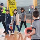 Em um ginásio de esportes, há equipamentos que simulam uma faixa de pedestres e um semáforo. No local, três meninas com cerca de 9 anos atravessam a faixa, orientadas por um homem com uniforme de agente de trânsito. Perto delas, há um menino segurando um volante