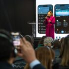Mulher vestida com um sobretudo rosa fala no microfone em um palco. Na plateia, há varias pessoas de costas, entre elas, um homem que usa o celular para tirar uma foto da apresentação