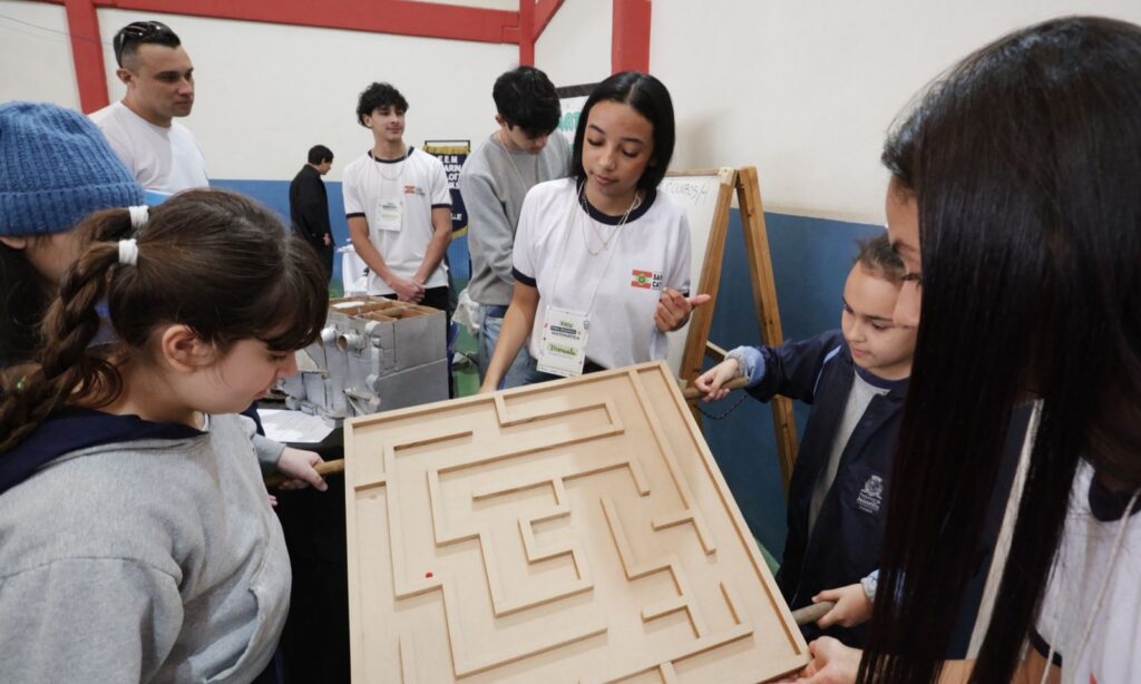 Duas meninas seguram um objetivo de madeira que simula um labirinto por onde passa uma pequena bolinha. Elas tentam equilibrar a bola no local e são observadas por outras estudantes