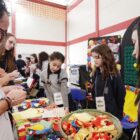 Estudantes apresentam um projeto de matemática envolvendo fuxicos. Há pedaços de tecido e fuxicos na mesa e pessoas adultas observam a explicação