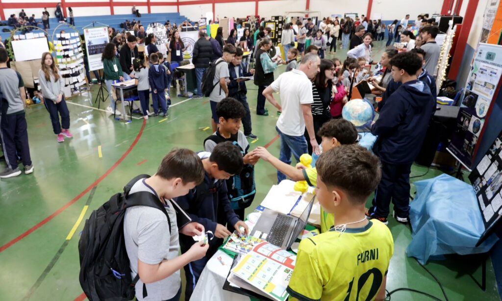Em uma quadra esportiva, há várias pessoas, a maioria jovens estudantes, observando trabalhos de matemática