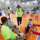 Imagem mostra duas crianças e um adulto sentados em cadeiras de roda, e outro jovem em pé próximo a eles. Um dos meninos está com uma bola de basquete e sorri