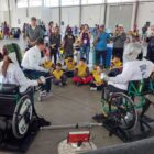 Imagem mostra adolescentes em cadeiras de roda utilizando espadas de esgrima. Ao redor deles há crianças e adultos sentados e em pé