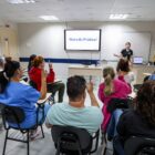 Em uma sala ampla, uma mulher jovem está à frente da sala, perto de uma televisão e de uma mesa. Há dezenas de homens e mulheres fazendo gestos em libras