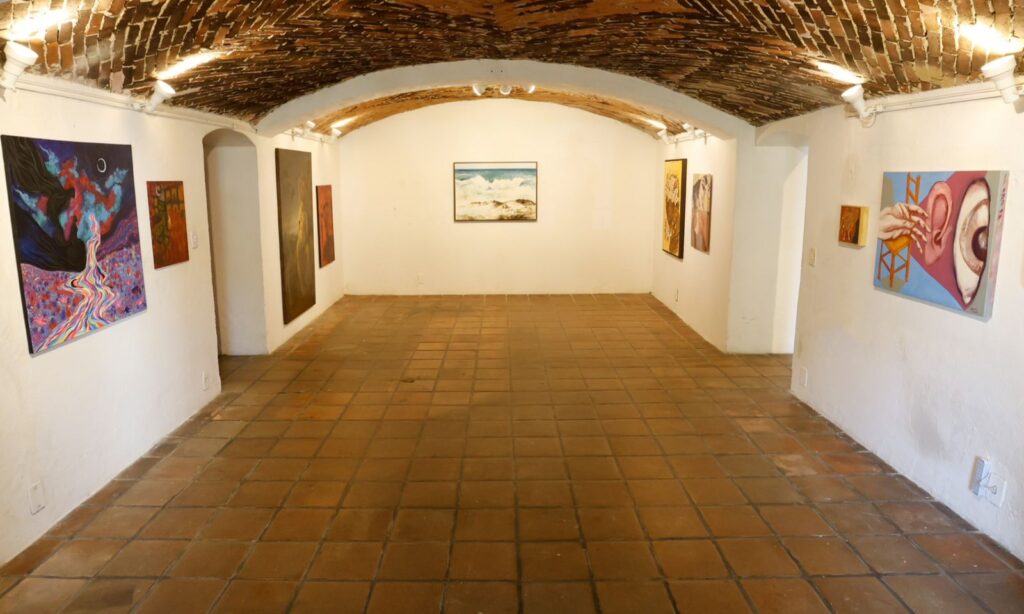 Imagem mostra o portão do Museu de Arte de Joinville, uma sala com forro em forma de arco e quadros pendurados nas paredes brancas