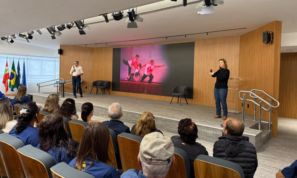 Em um palco, há um homem falando no microfone à esquerda, um grande telão com uma imagem de dança no centro e uma mulher fazendo libras à direita. Na plateia, há homens e mulheres sentados de costas para a câmera.