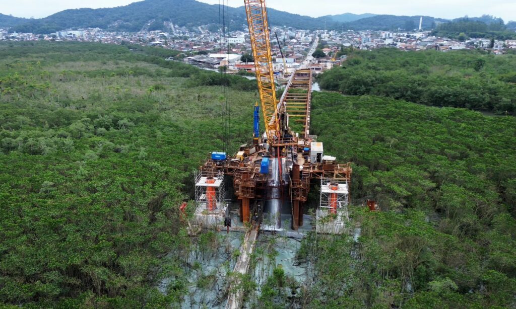 Imagem mostra escavação de estacas na Ponte Joinville. A imagem aérea mostra uma área com ferragens, máquinas e outros equipamentos