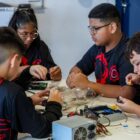 Três meninos e uma menina estão uniformizados com camisetas pretas e detalhes em vermelho. Eles estão em uma mesa, onde manuseiam materiais de robótica